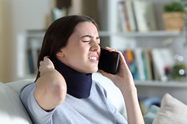 Woman with a neck brace talking on the phone with the insurance company after a car accident while sitting on a couch at home.
