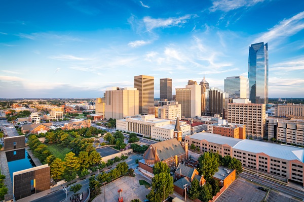 Aerial view of downtown Oklahoma City with modern skyscrapers, historic buildings, and tree-lined streets under a clear evening sky.