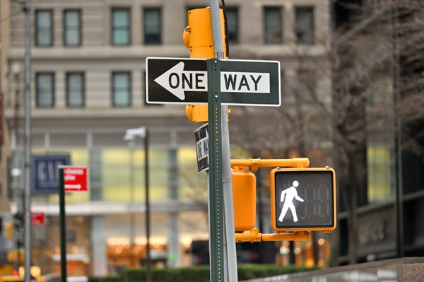 Pedestrian crosswalk signal showing a walk symbol next to a one-way street sign in a city.