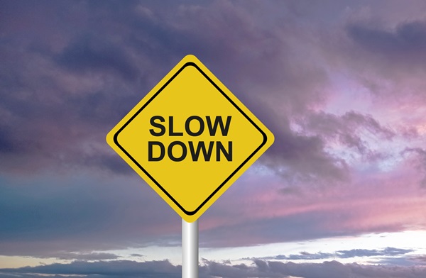 Here is the alt text for the image: A bright yellow, diamond-shaped warning sign on a pole that reads "SLOW DOWN" in black letters, set against a dramatic, cloudy purple and pink sunset sky.