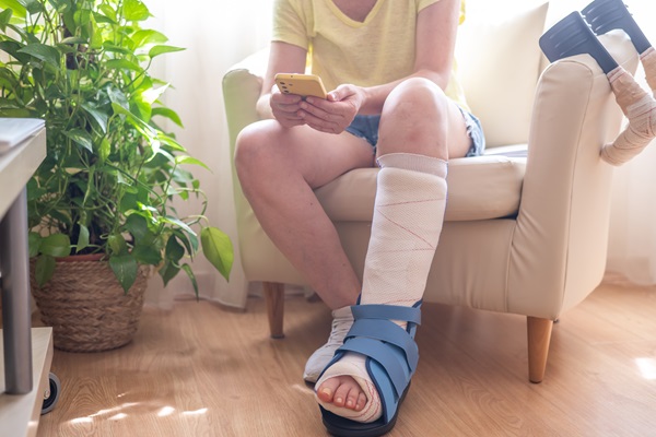  A person sits in a chair using a smartphone while wearing a large medical walking boot and cast on their leg.