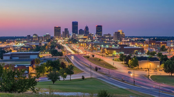 A wide-angle, twilight view of the Oklahoma City skyline with long-exposure light trails from traffic moving along the highway in the foreground.