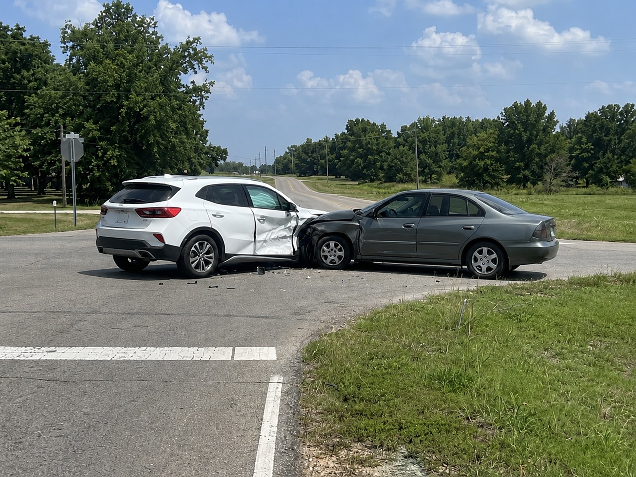 A gray sedan has T-boned a white SUV at a rural Oklahoma intersection, leaving the SUV with severe rear quarter panel and door damage and debris scattered across the pavement, depicting a failure-to-yield crash where establishing which driver had the right of way is central to proving fault and recovering compensation under Oklahoma law.
